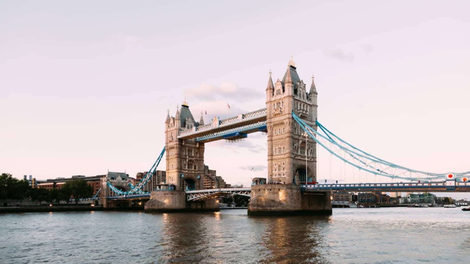 The iconic Tower Bridge of London spanning the dark Thames River in the foreground against a pale, dusky sky.