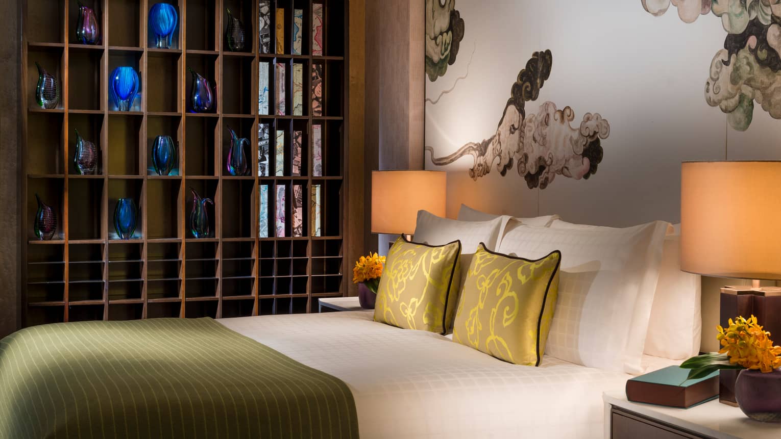 Hotel bed with gold silk pillows under wall shelf lined with blue, purple vases