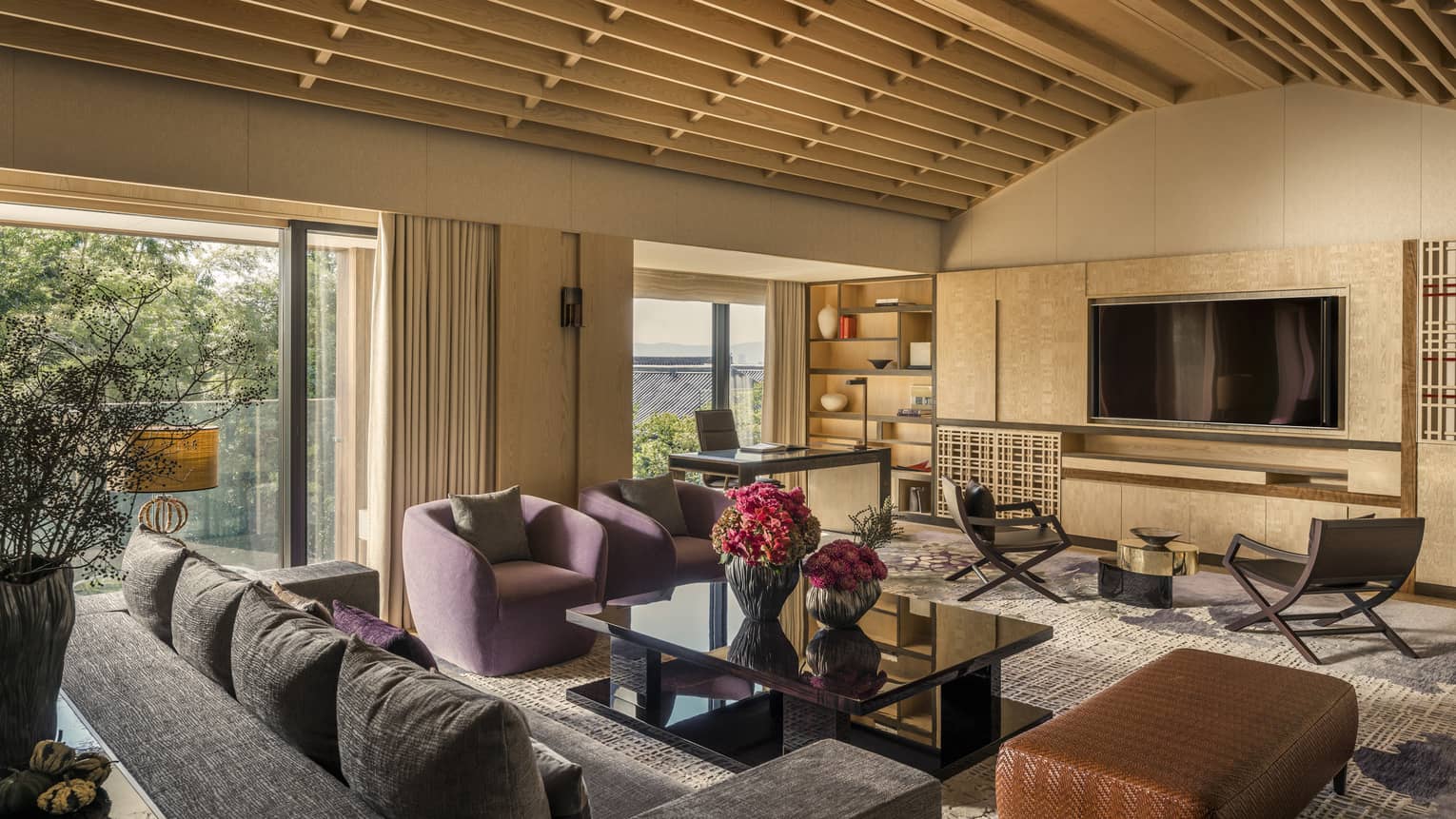 Living area with vaulted ceiling, purple armchairs, sofa, black coffee table, wall-mounted TV, desk, chairs and large windows overlooking greenery