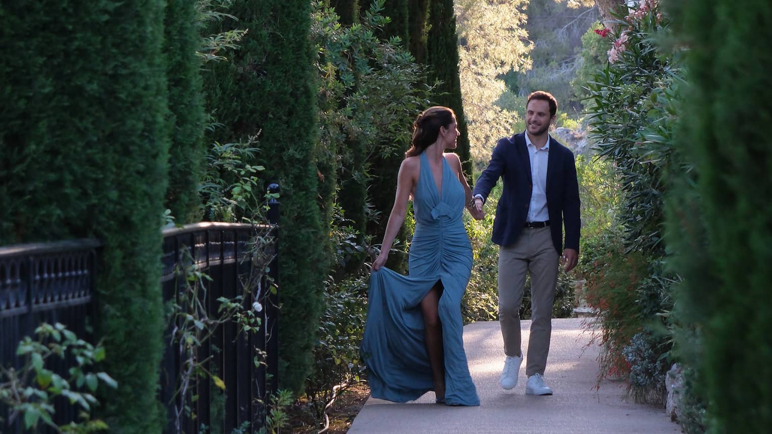 A couple dressed in formal attire walks along a shaded pathway surrounded by tall, lush trees