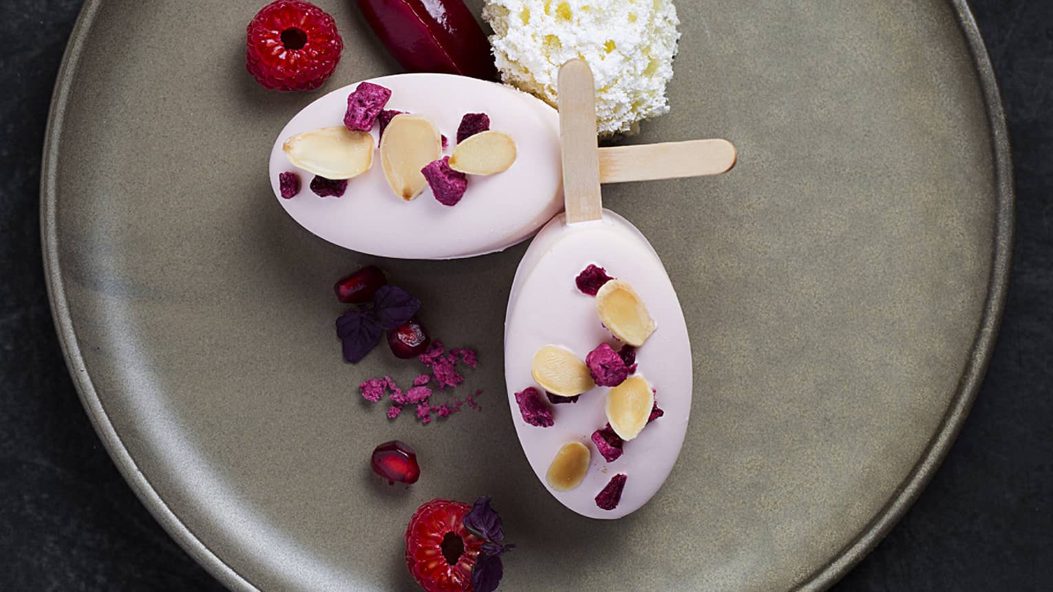 Two Galangal Ginger Soya Milk Ice Cream popsicles on a gray stone plate garnished with raspberries and red and yellow flower petals  