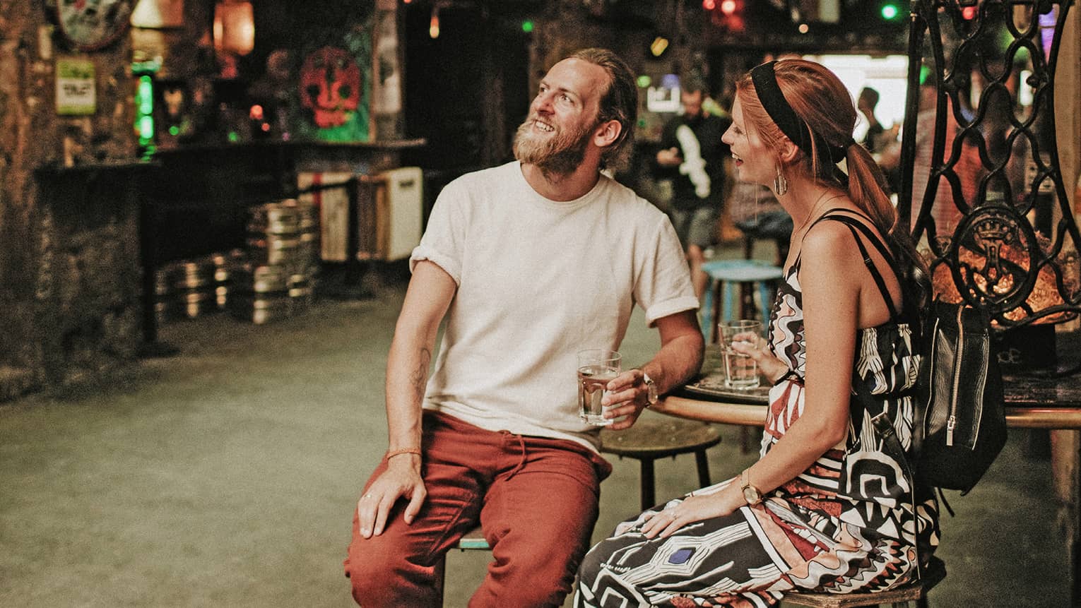 A smiling couple holding drinks, surrounded by myriad disparate objects and scrap parts on the walls and hanging overhead.