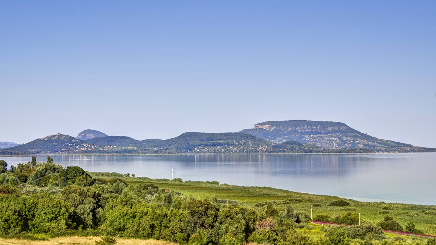 A clear, sunny day at a lake surrounded by lush green vegetation; across the lake is a volcanic hill with homes built on it.