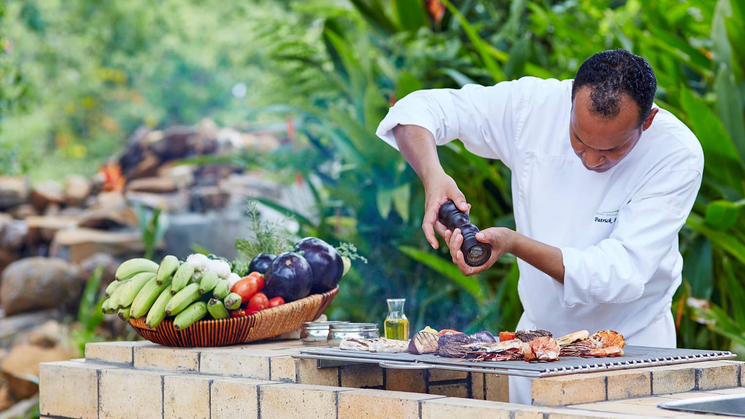 Close-up of chef grinding pepper over meat on grill by basket of fresh fruits and vegetables