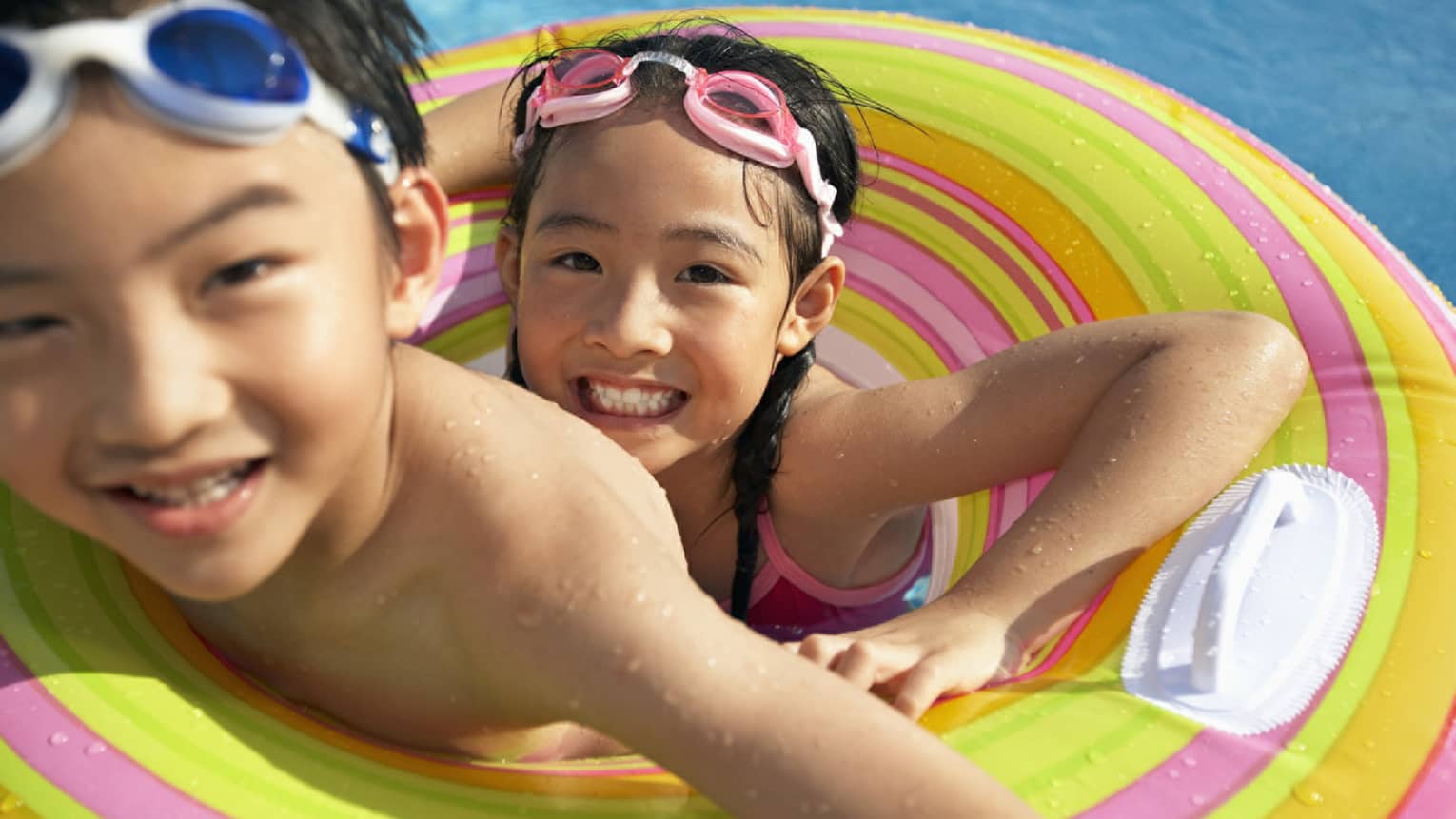 Smiling brother and sister with goggles on head share inflatable tube in swimming pool