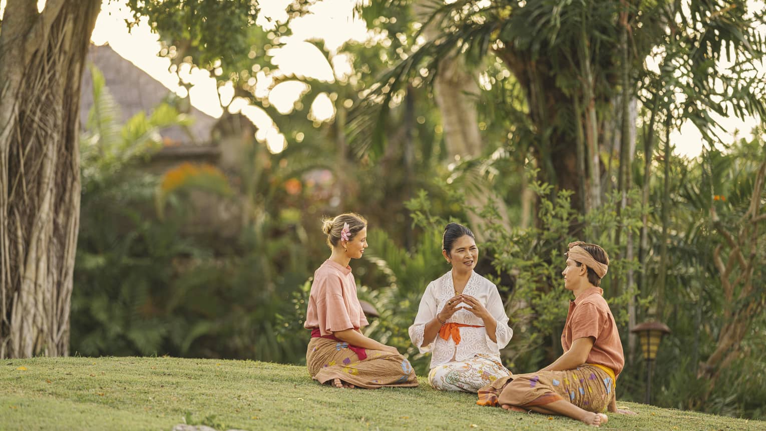 Two guests in sarongs listen to an elder as they all sit on a grassy knoll surrounded by lush greenery and palm trees.