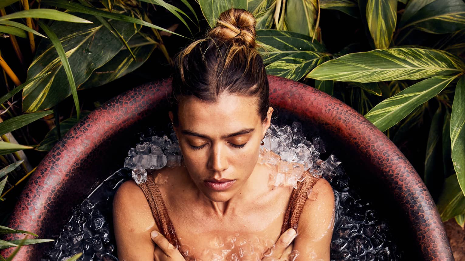 A guest, wearing a bathing suit, is sitting and relaxing in a rustic bathtub full of ice with foliage encircling the tub.