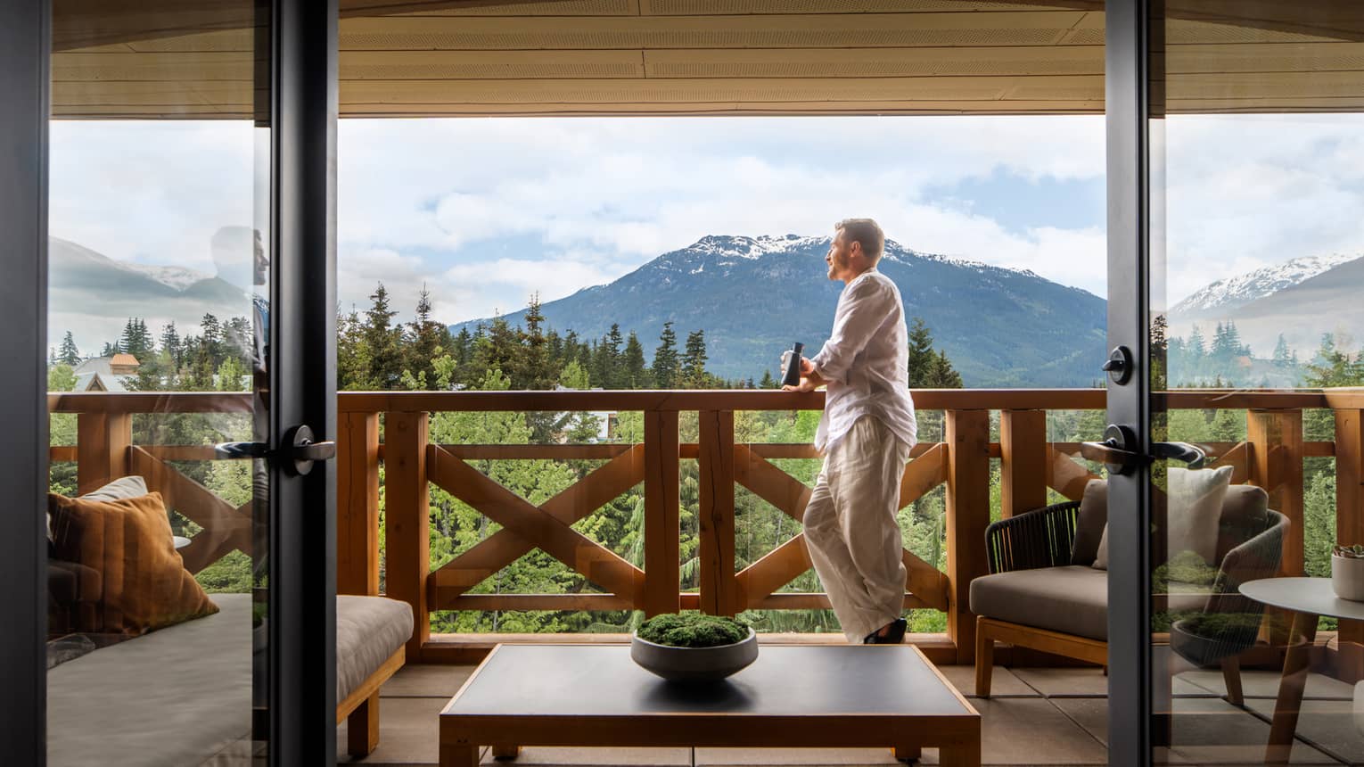 Man with short hair wearing a white long-sleeve shirt and khaki pants leans on the railing of a balcony with a beer in hand, looking out onto the mountain views
