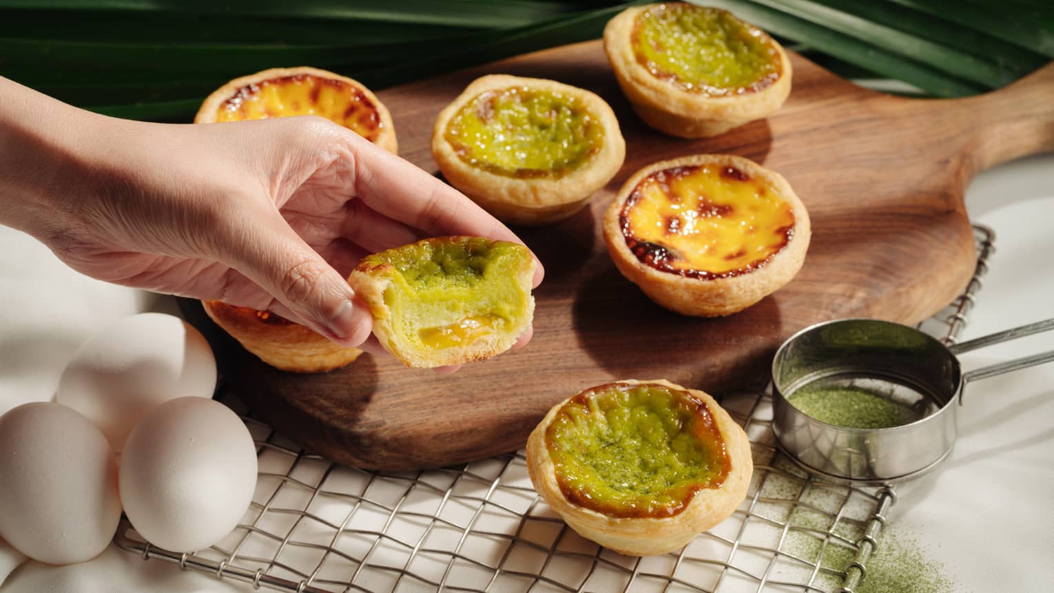 A hand holding a half-eaten Portuguese egg tart with a green filling, set against a backdrop of whole and partially-eaten egg tarts on a wooden board.