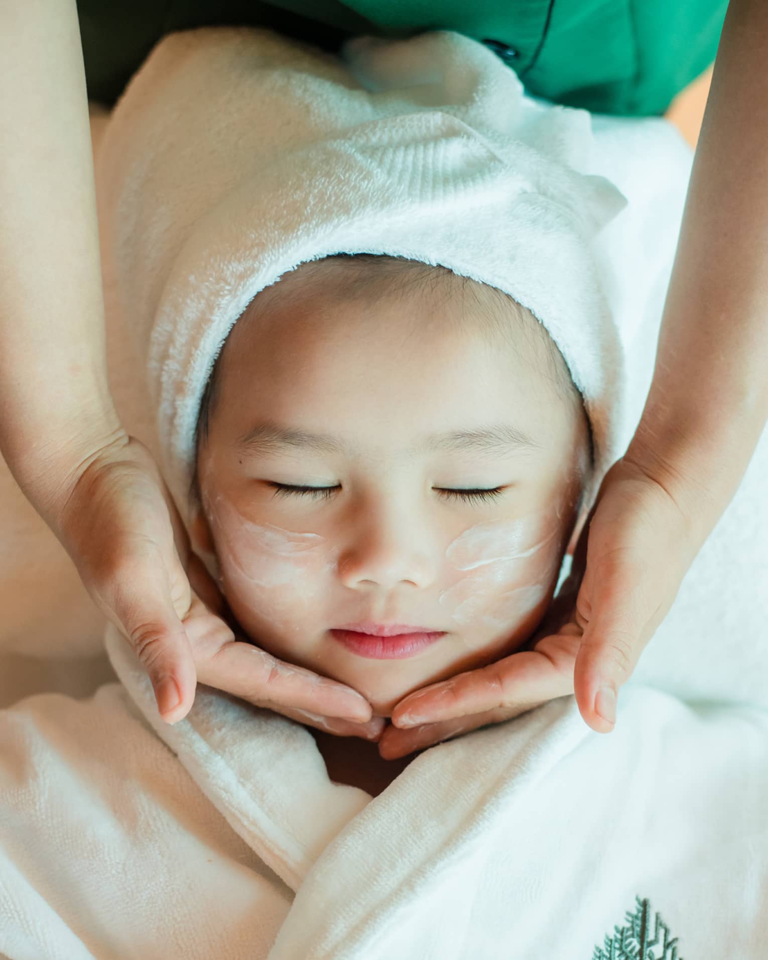 A close-up of a relaxed child wrapped in a towel and robe and enjoying a masseuse massaging a cream into their face.
