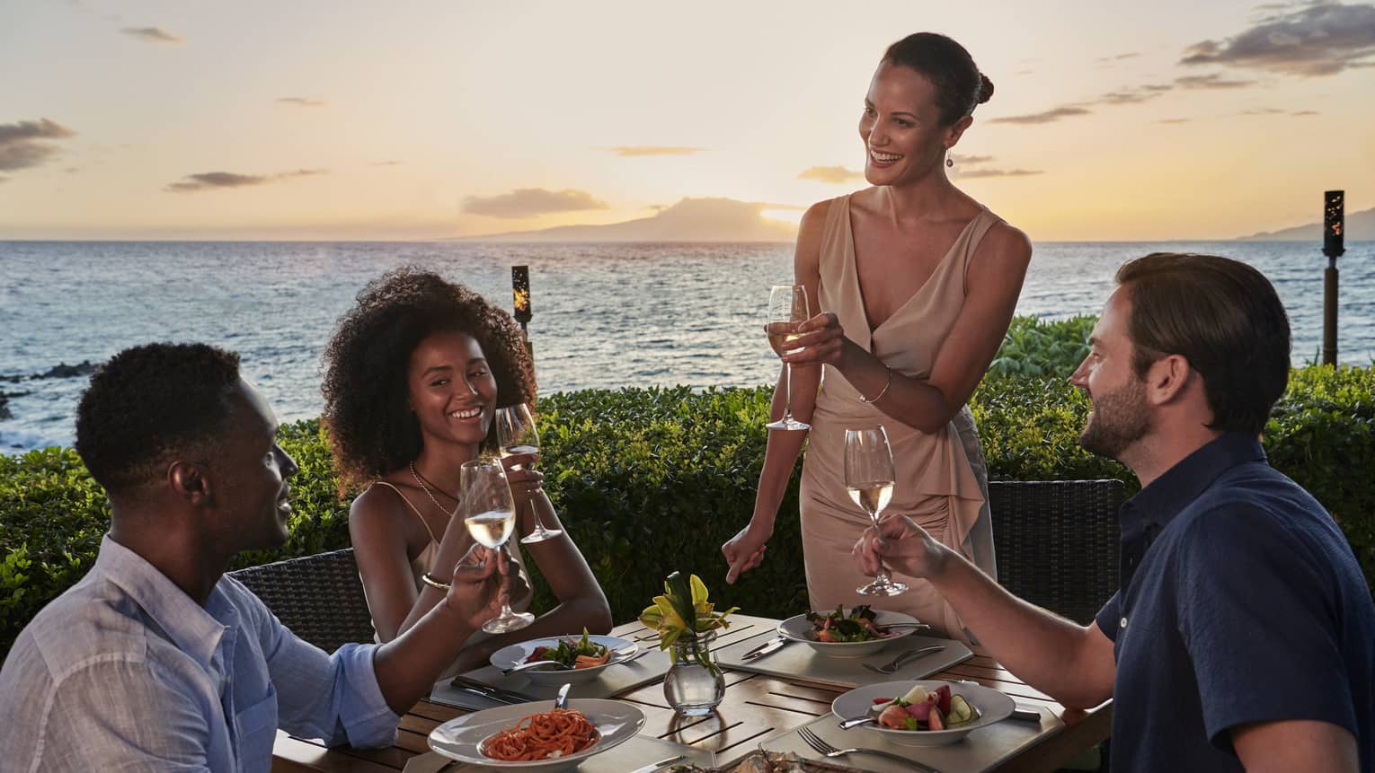 Two women and two men toast wine glasses at outdoor, oceanfront restaurant