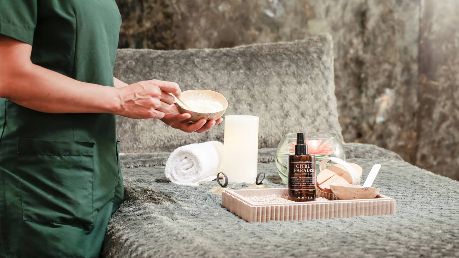 Body view of spa attendant mixing salt scrub in small bowl over bed with tray, towels, lotion