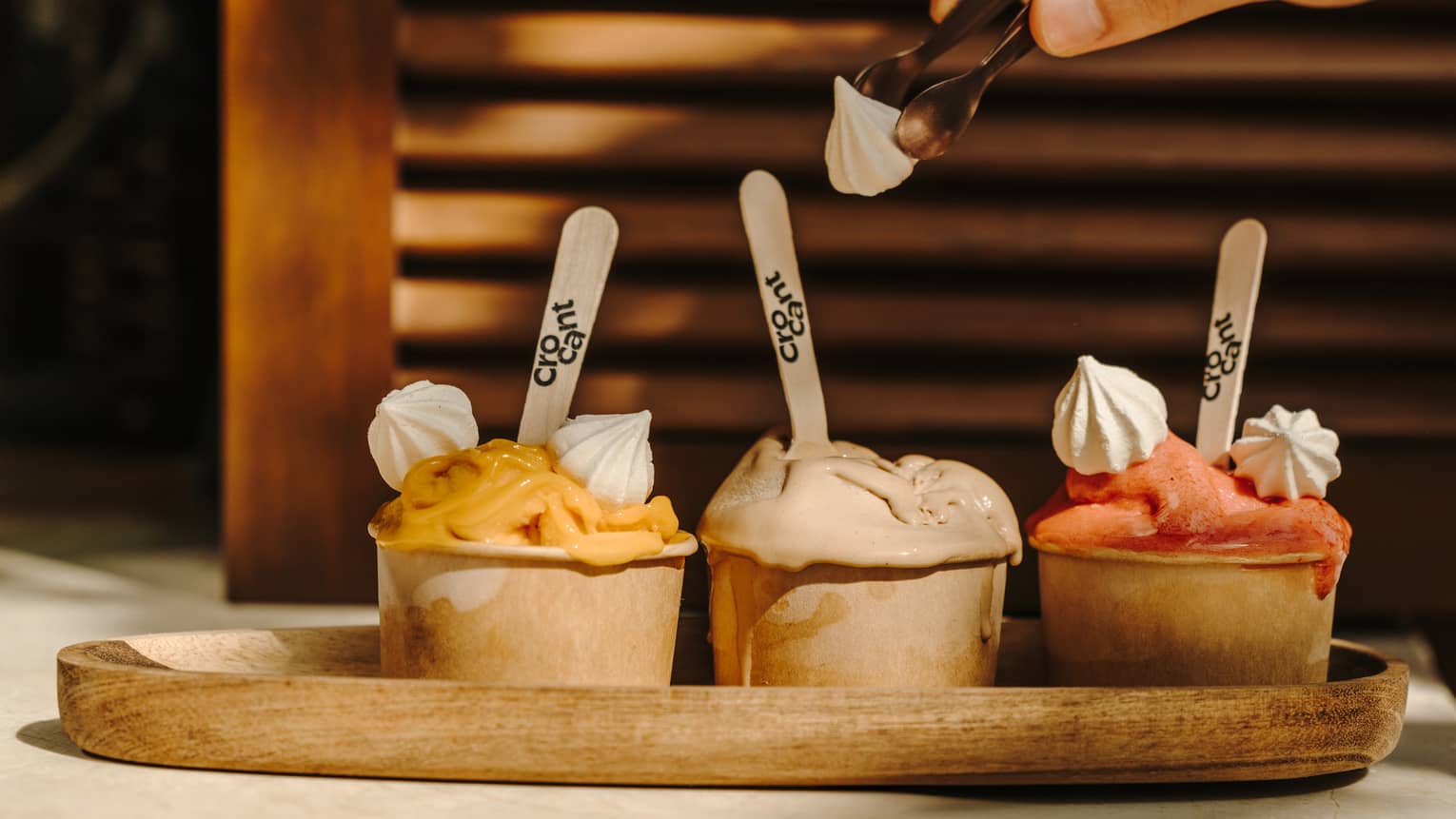 A person adds toppings to three cups of ice cream on a wood serving plate.