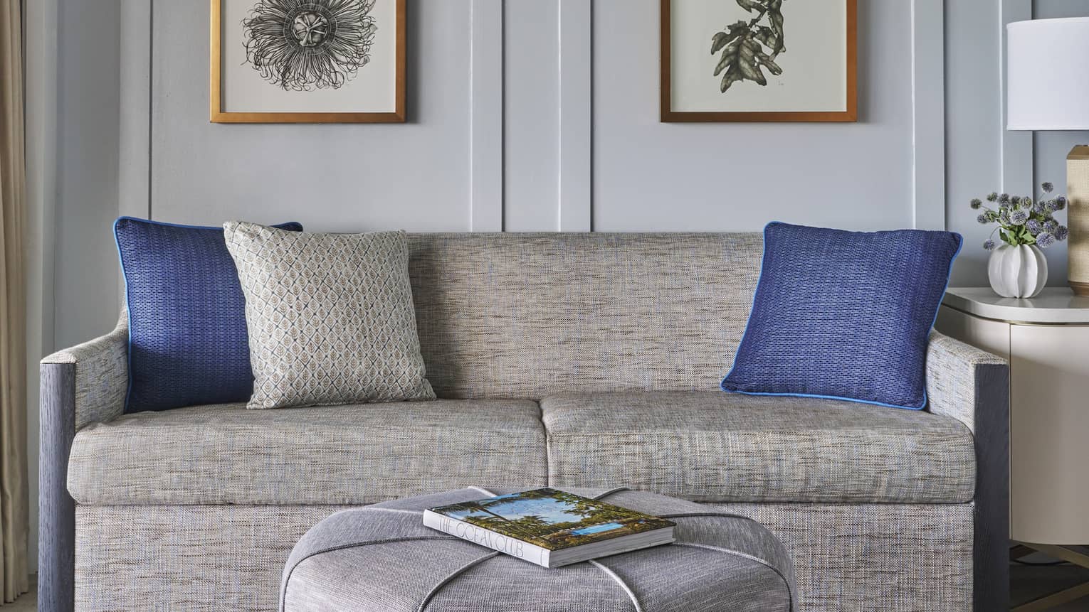 Two-seat sofa and grey, round ottoman/table, set against a wall with four hanging flower pictures
