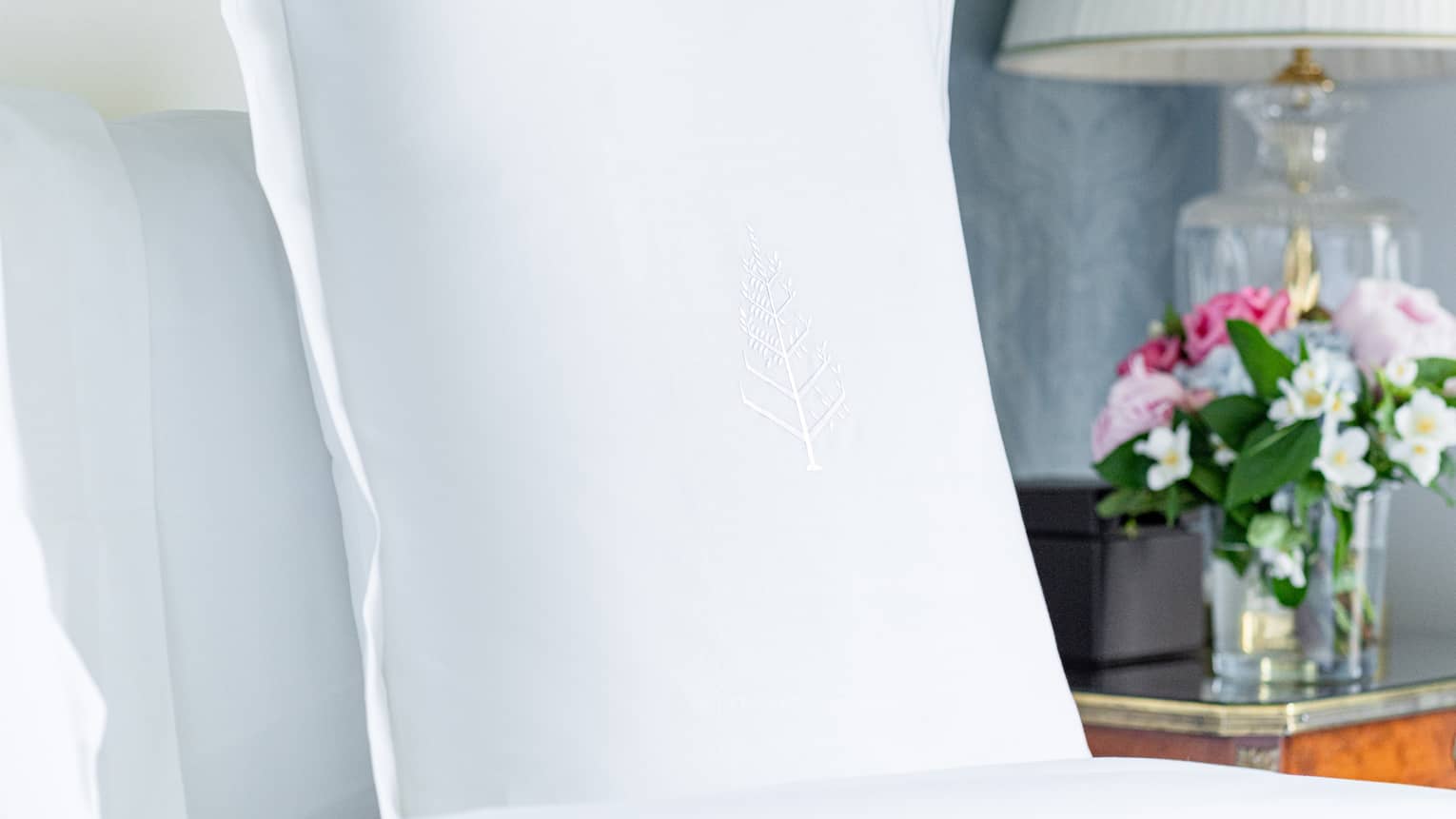 Close-up of a bed pillow with an embroidered Four Seasons logo and a bedside table with a floral arrangement and lamp