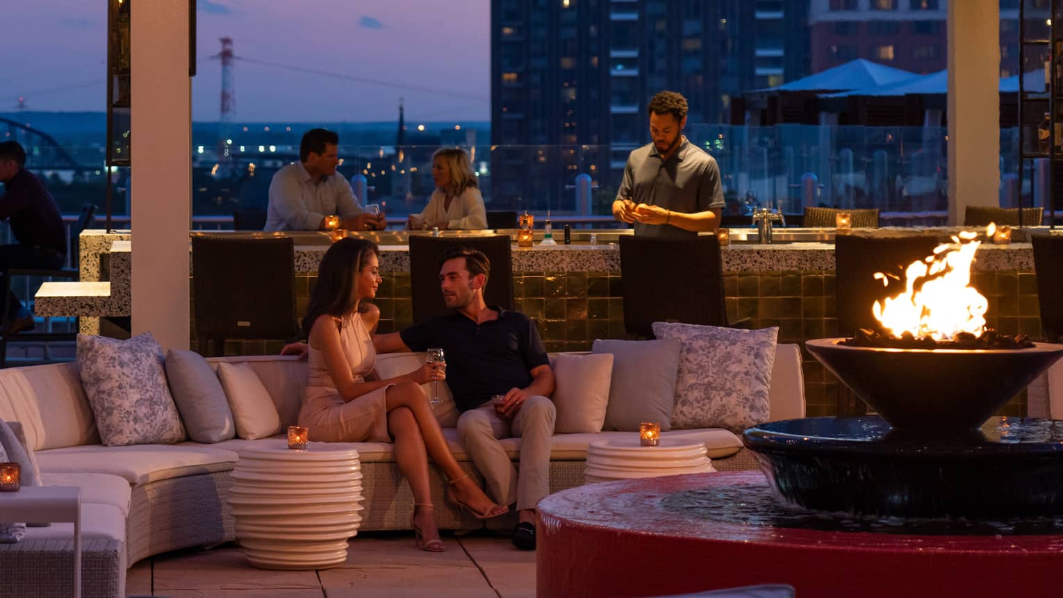 The sky is dark beyond the cinderhouse rooftop bar – furnished with a curved sectional, fire pit, hanging lights and people socialize 