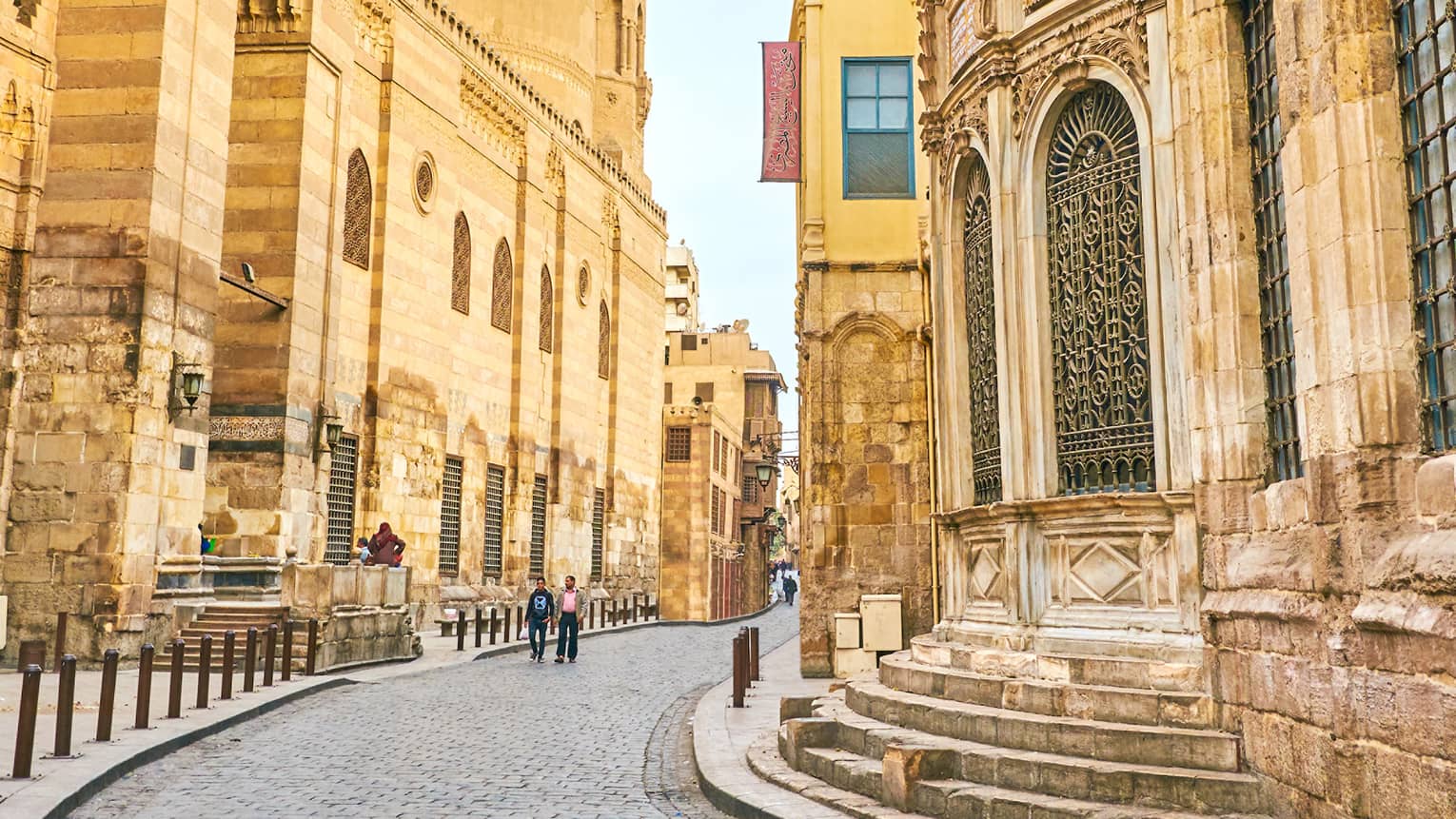 Pedestrians walk along a meandering cobbled road between tall, yellow stone buildings with ornate lattice windows.