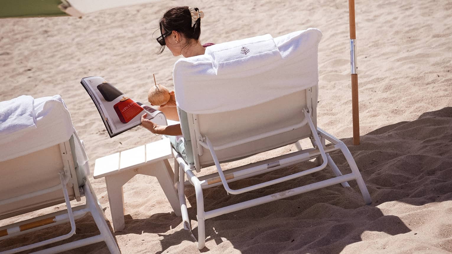 A woman lounges on a beach chair with a cocktail underneath a green and white umbrella