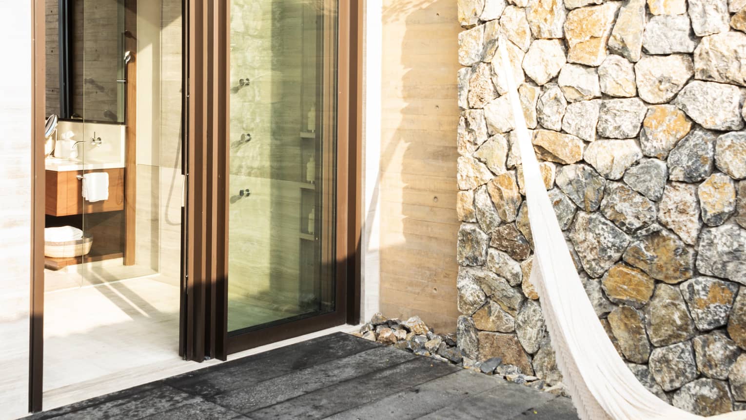 Outdoor patio with stone walls, a hammock and large terracotta pots. Glass doors lead to a bathroom with a shower, blending indoor and outdoor living.