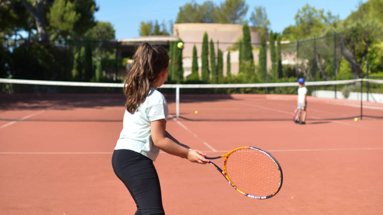 Girl with brown ponytail in light blue T-shirt and black pants playing tennis on clay court