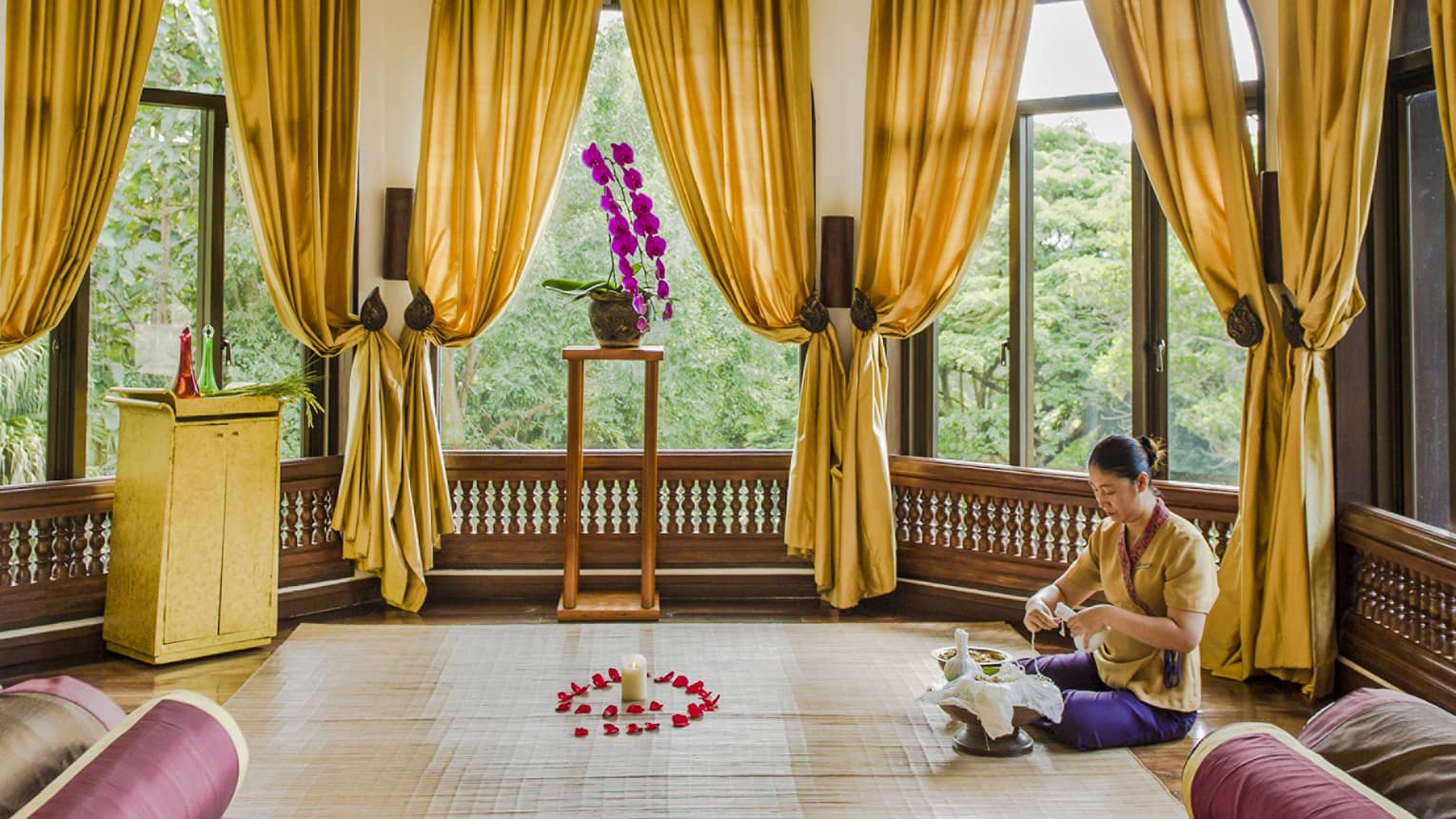 A person meditating in a window filled room with golden curtains, a red chaise, and golden embellishments on the ceiling.
