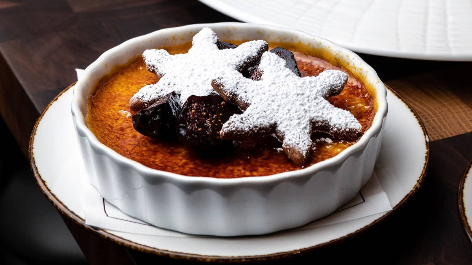 Vanilla Cr�me Br�l�e in a round white dish topped with figs and powdered-sugar-covered snowflake cookies