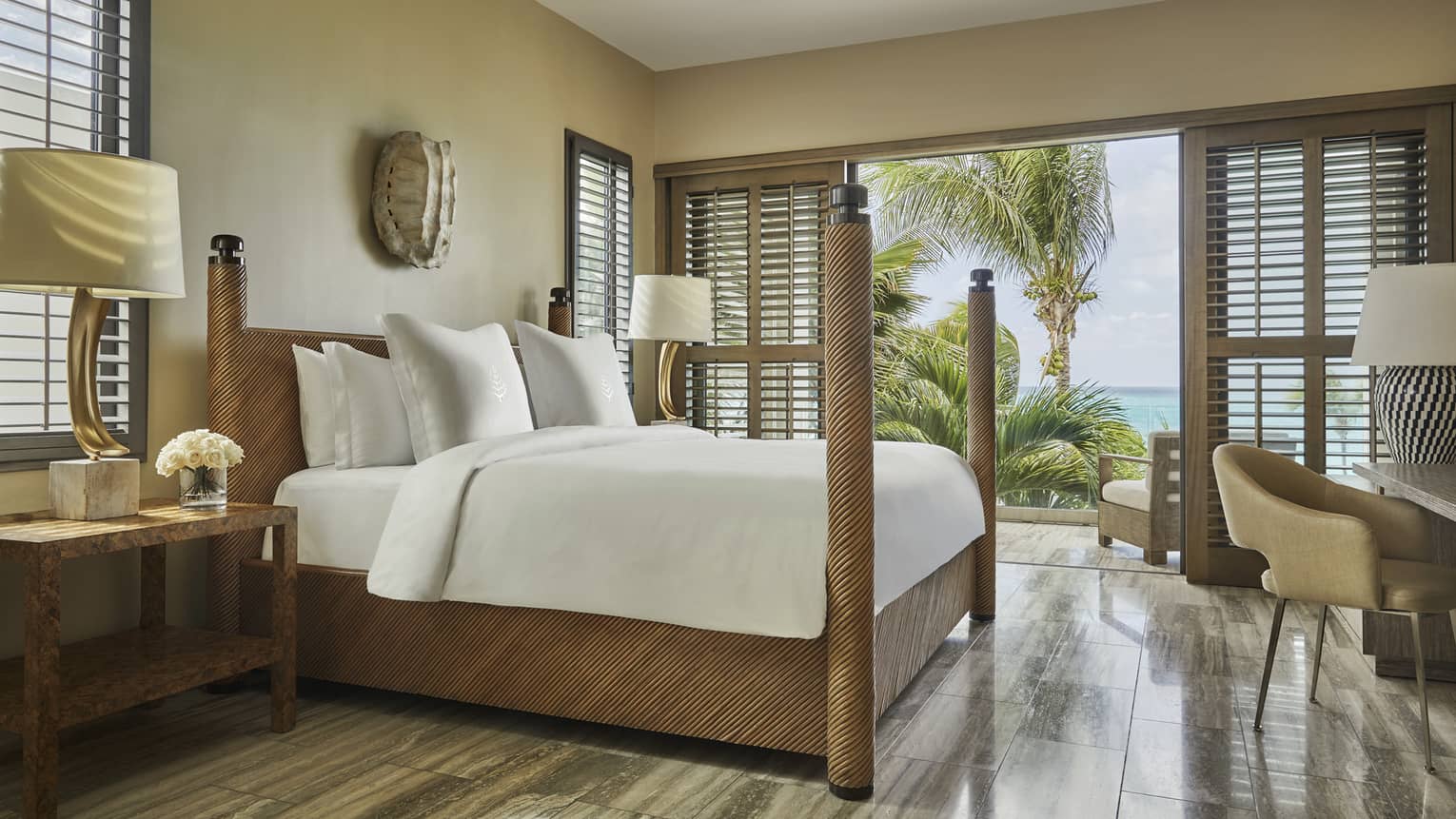 Luxury beachfront villa bedroom with a four-poster bed, white bedding and decorative lamps, featuring a balcony view of tropical palm trees and the ocean
