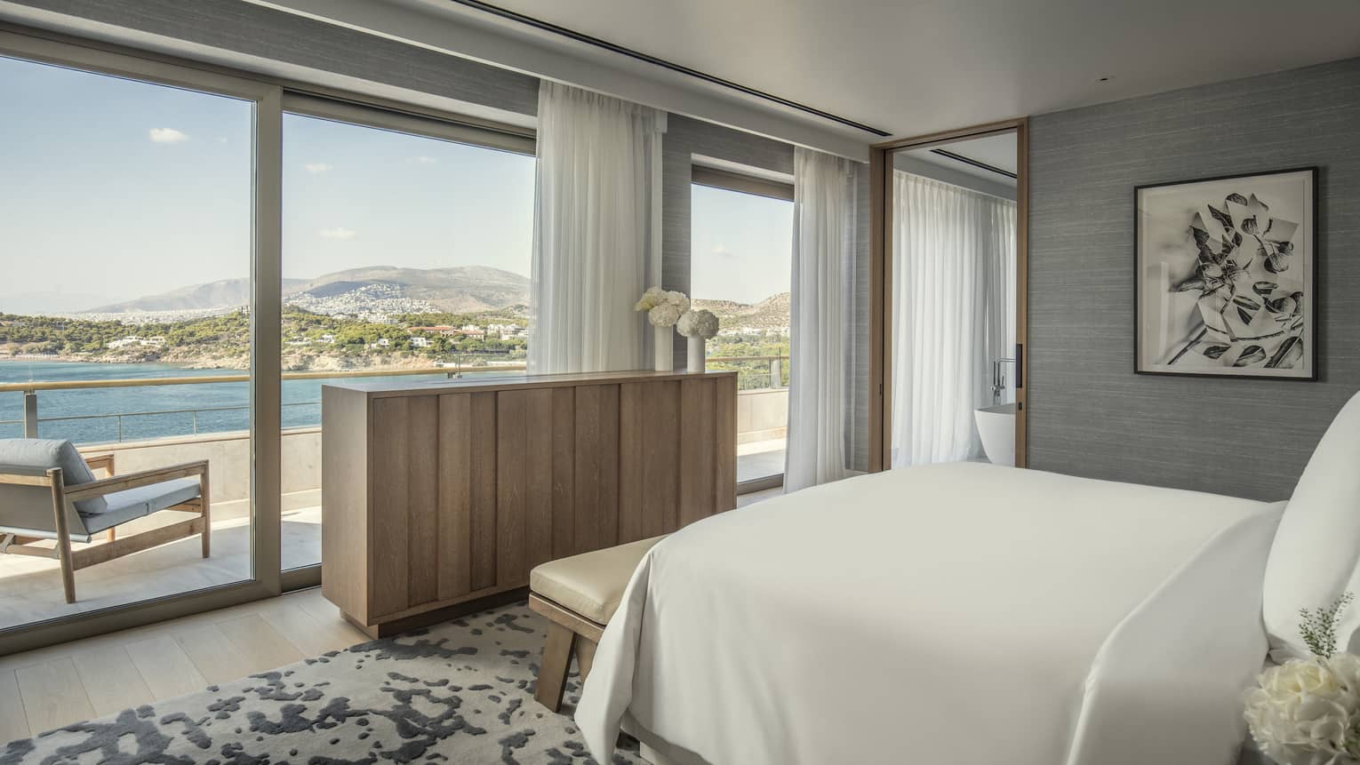 Arion Presidential Suite with white King bed, matching wall art and area rug, blonde wood sideboard, floor-to-ceiling window with ocean views