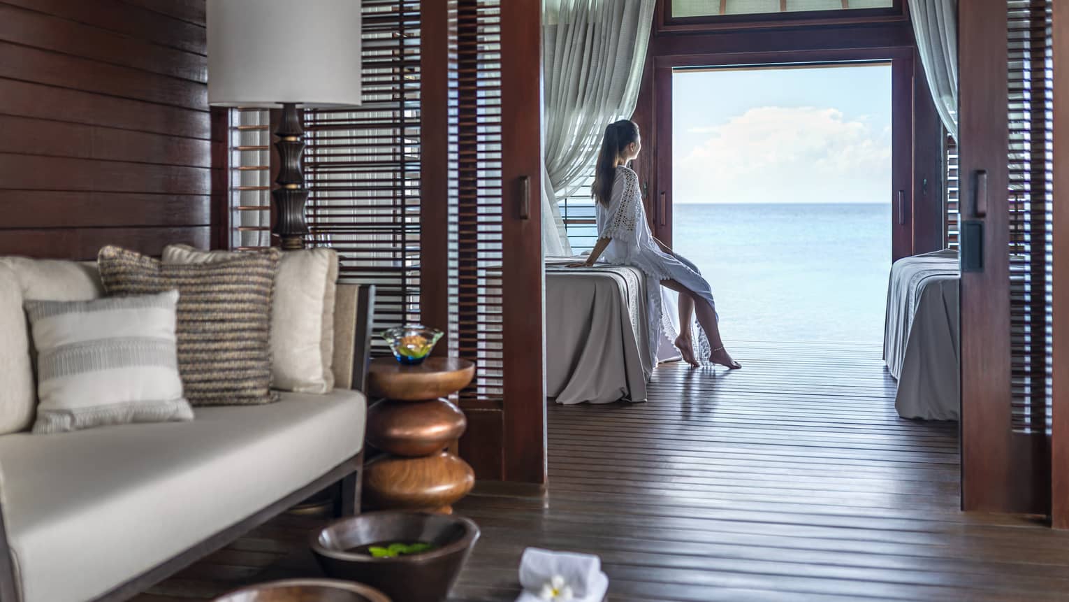 A woman leans on a massage bed on the far side of a spa treatment room and looks out through a large doorway onto the vast blue ocean