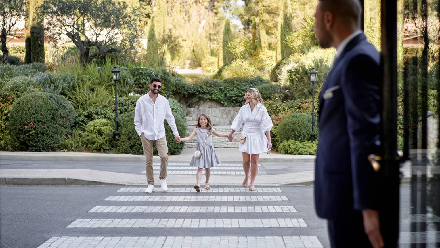 Couple and young girl hold hands, cross road toward hotel door