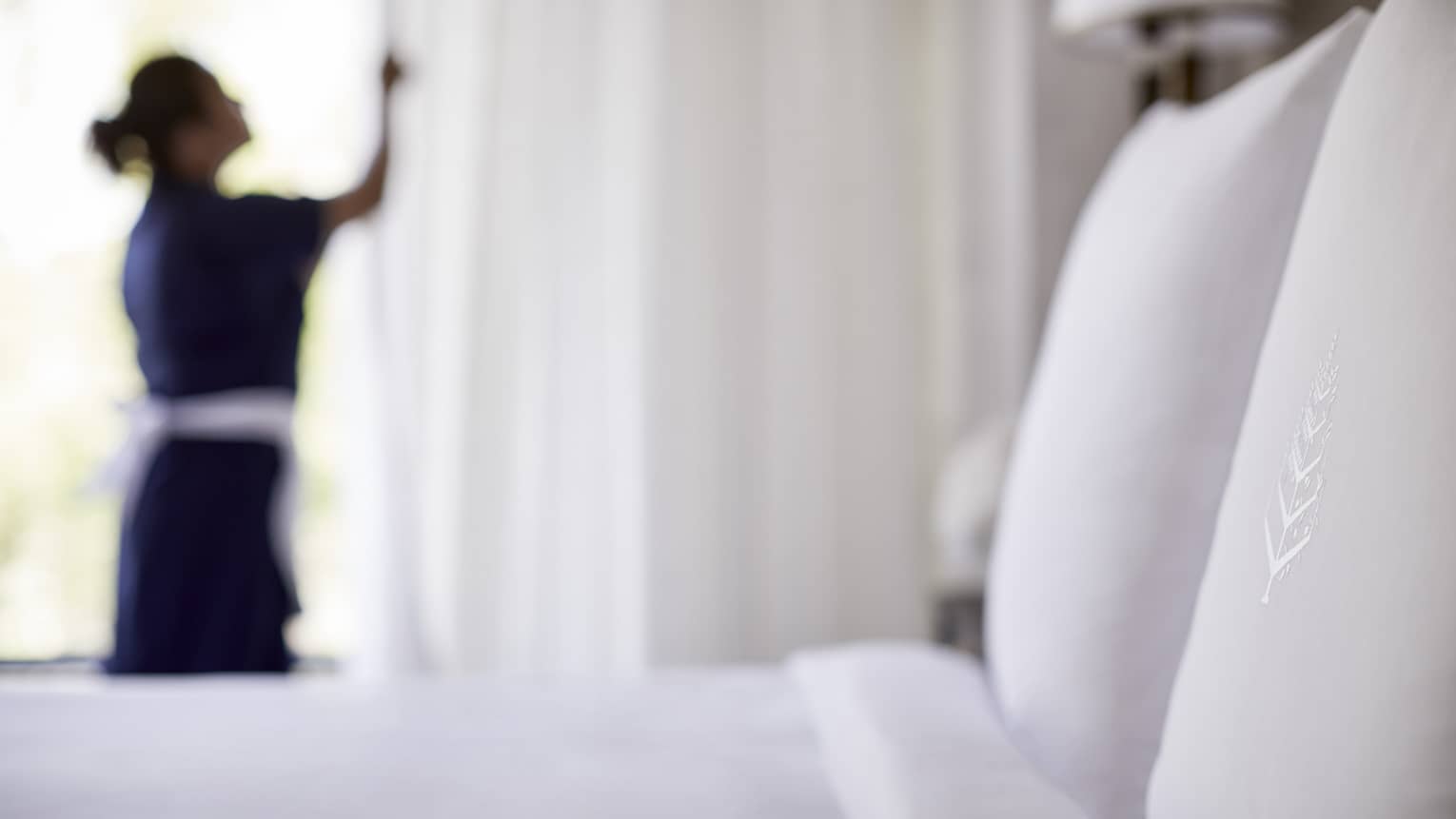 Close-up of bed, pillow with silhouette of hotel staff in uniform closing white curtains in background