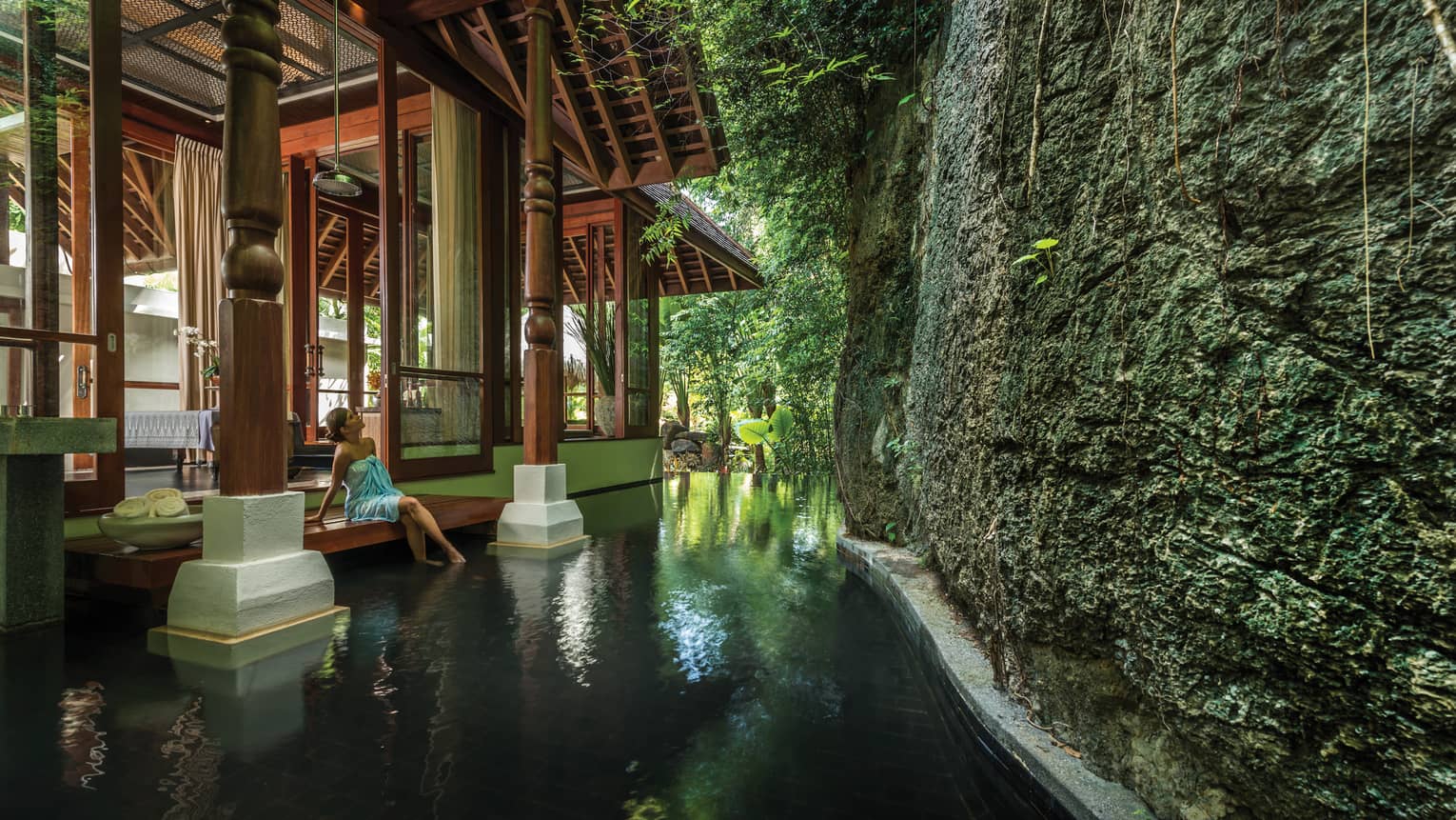A woman dips her feet in a pond while looking up at a mossy rock wall that encloses The Geo Spa at Four Seasons Resort Langkawi. 