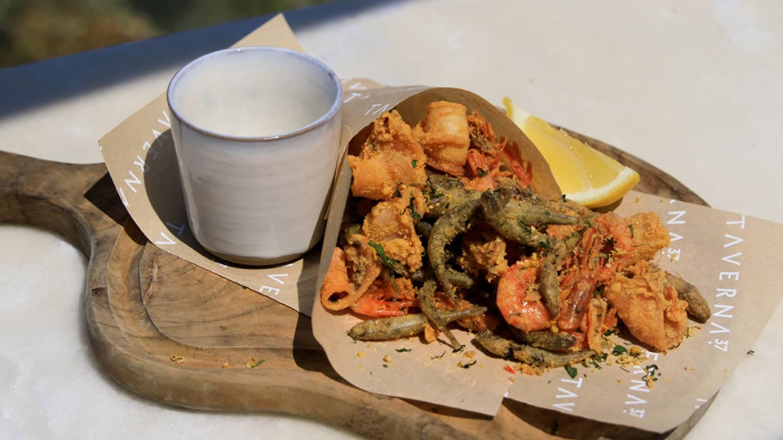 Fried seafood wrapped in paper on cutting board with cup of caper sauce and lemon wedge