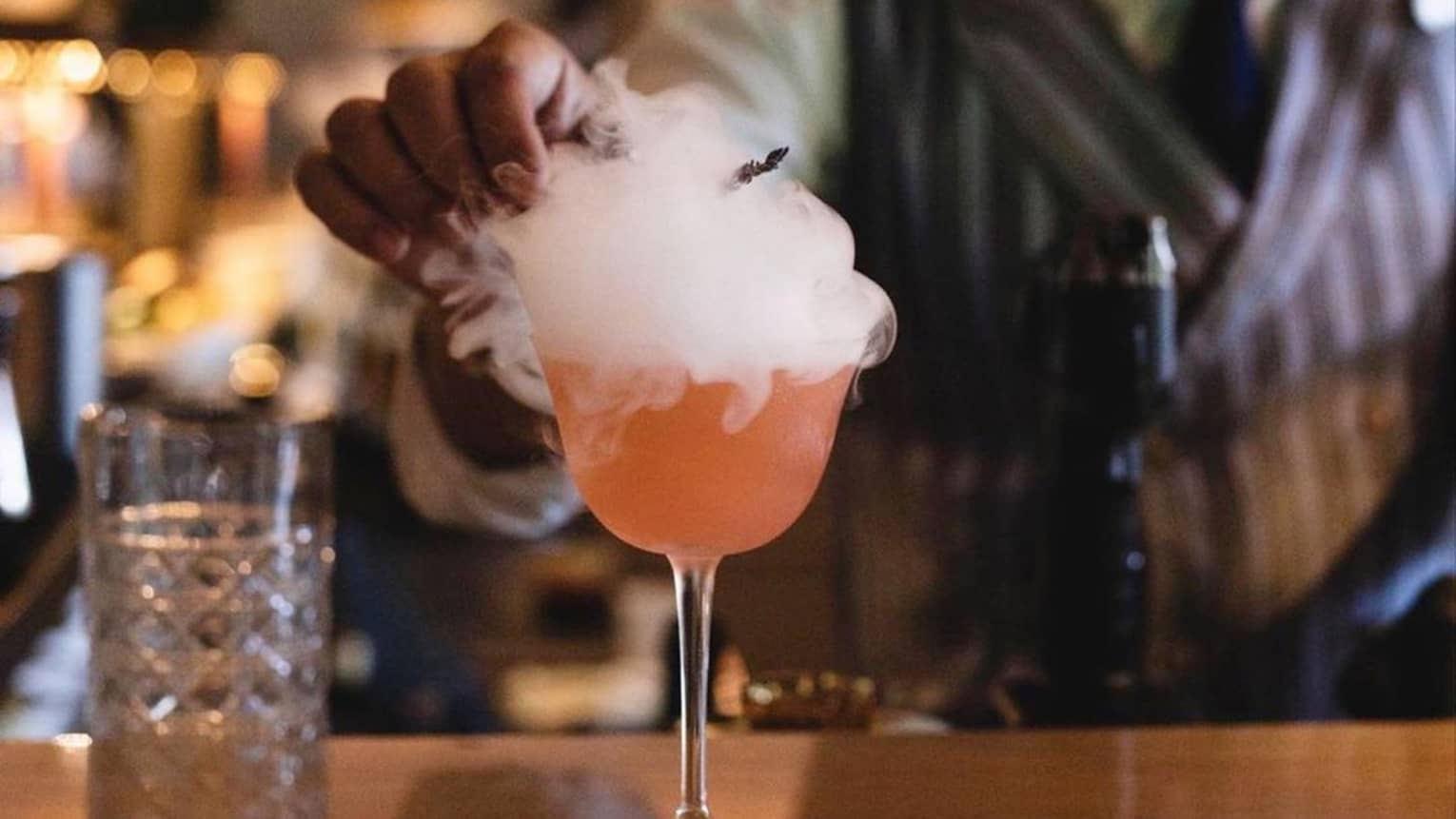 A mixologist garnishes a bright orange cocktail atop a smooth wooden bar; smoke billows over the sides of the stemmed glass.
