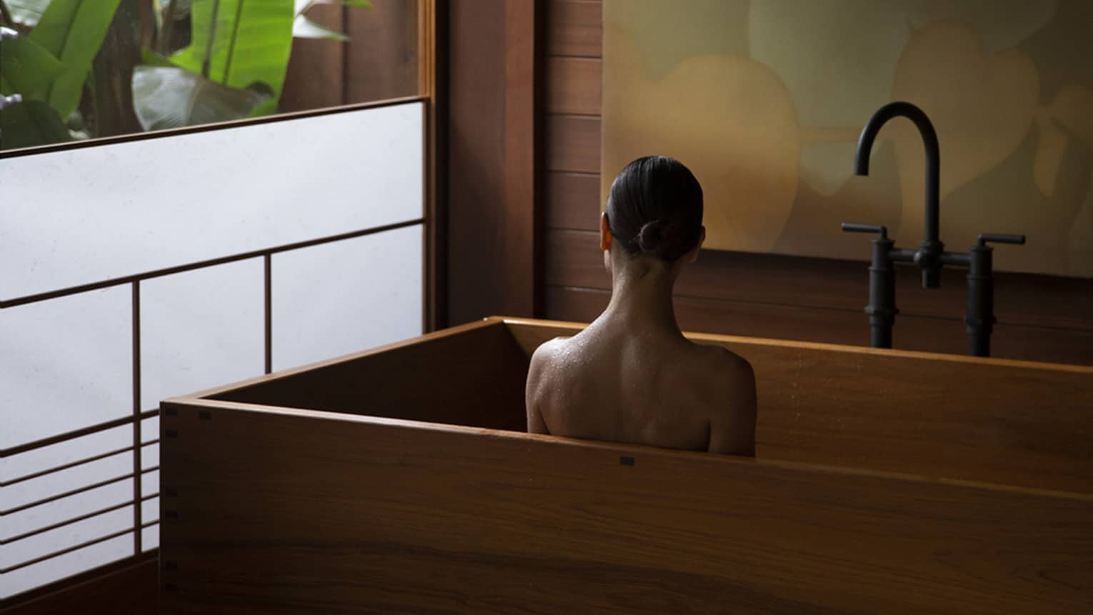Woman sits in wooden, modern tub next to semi-frosted window