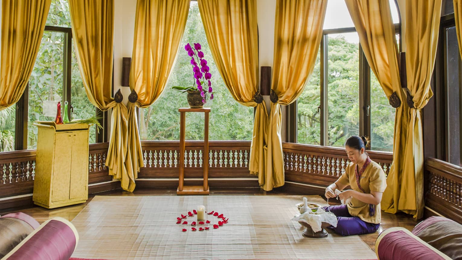 A person meditating in a window filled room with golden curtains, a red chaise, and golden embellishments on the ceiling.