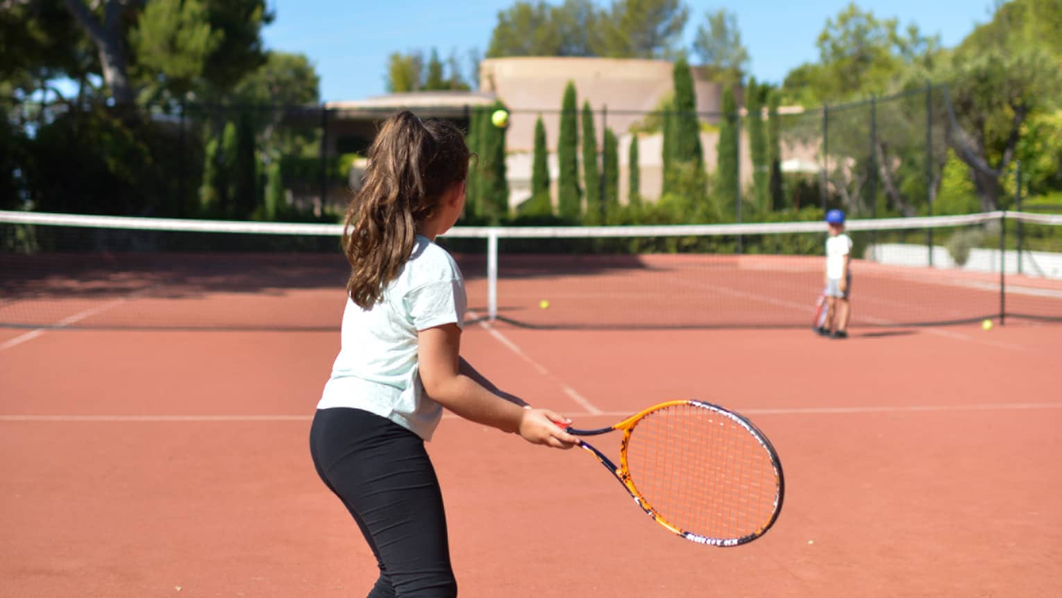 Girl with brown ponytail in light blue T-shirt and black pants playing tennis on clay court