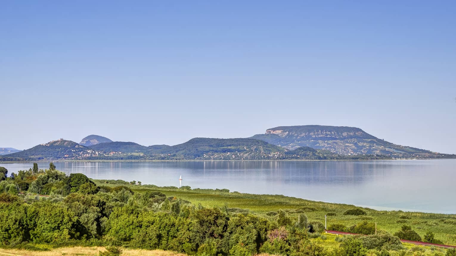 A clear, sunny day at a lake surrounded by lush green vegetation; across the lake is a volcanic hill with homes built on it.