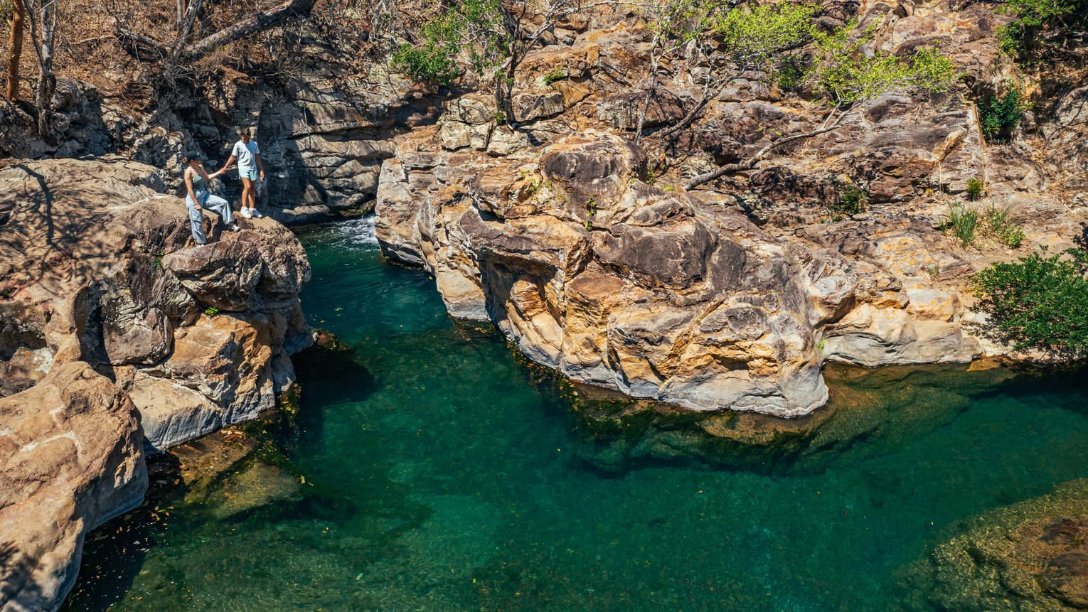 Two people stand holding hands at the edge of a rockface overlooking a blue-green lagoon below that flows through a rocky landscape