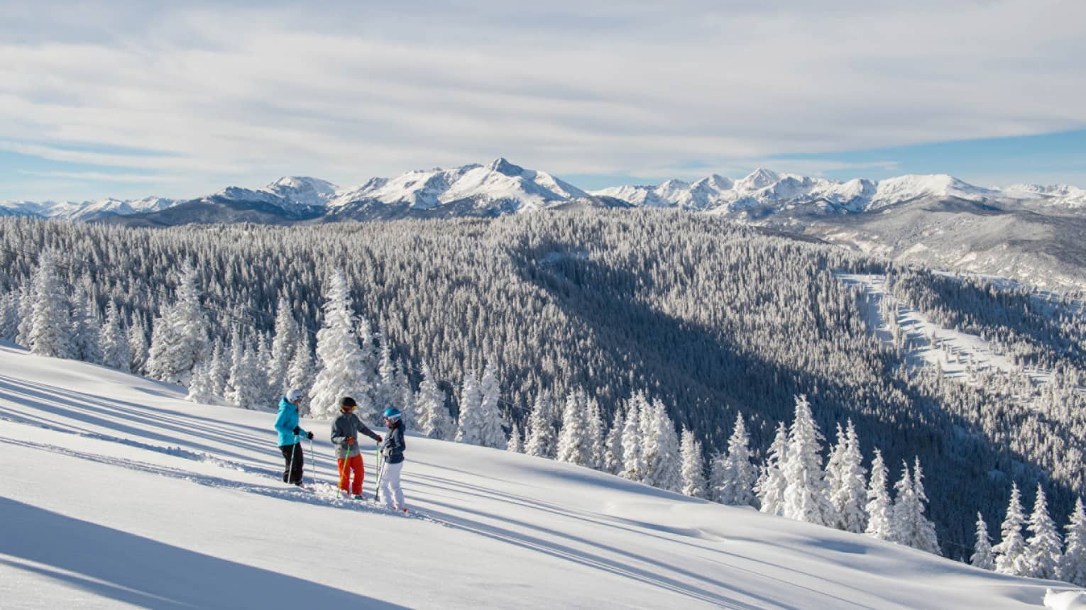 Three guests on skis pause on the ski hill, snow capped trees and mountains in distance 