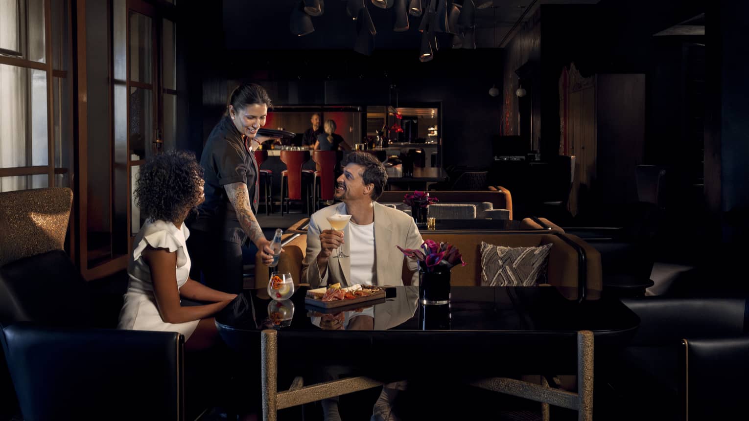 Two people wearing light-coloured cocktail attire sit on benches around a black table as a waitress brings them beverages