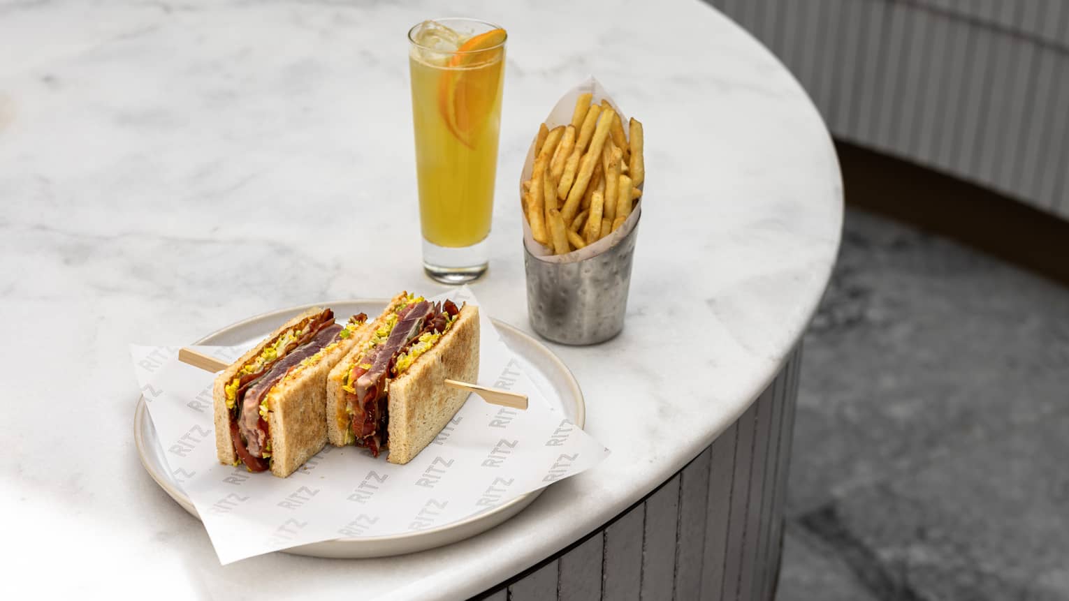 Two sandwiches on a plate next to a pale orange drink in a clear glass and a container of French fries, all on a white table
