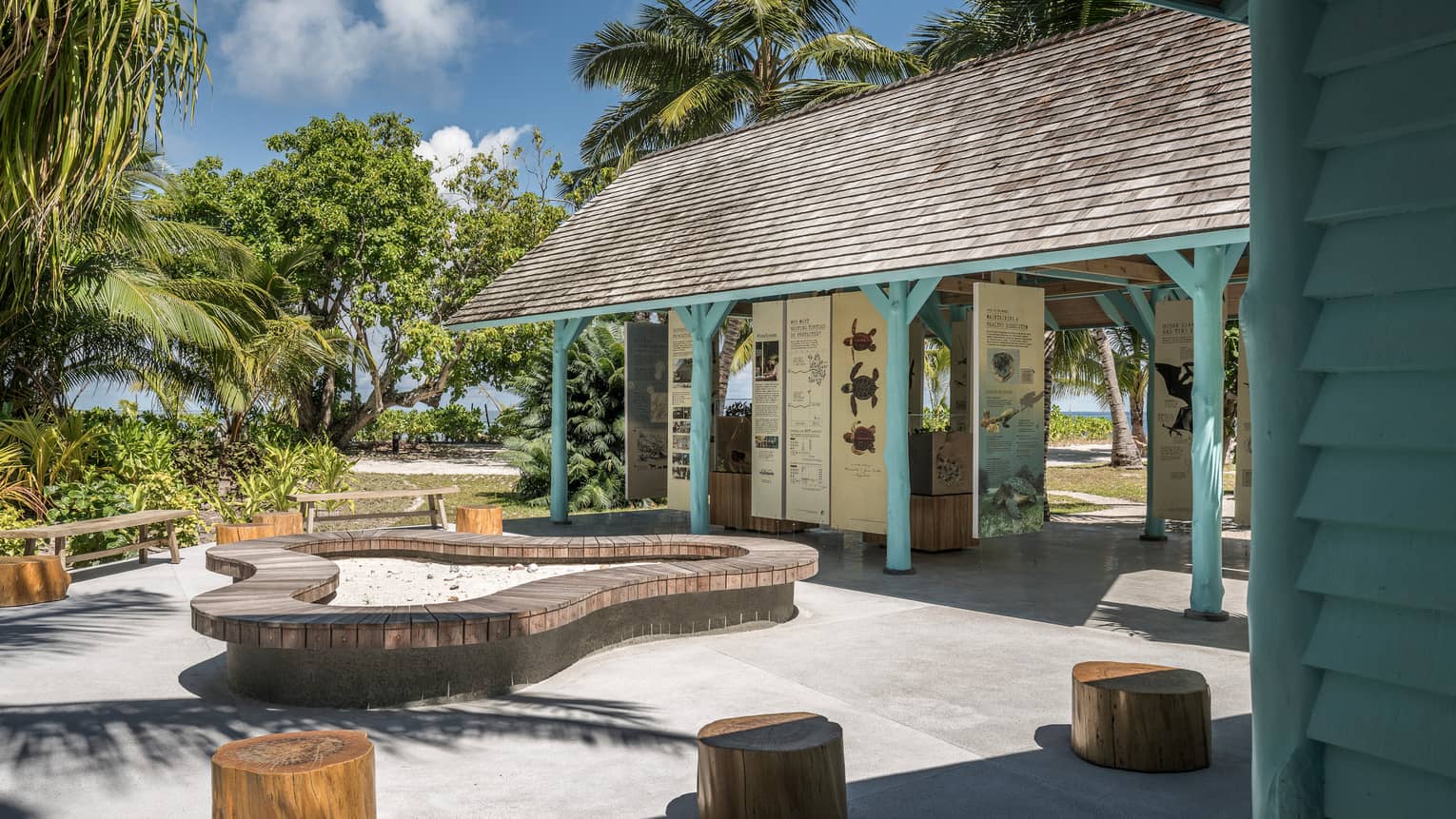 Outdoor information hut with blue poles, surrounded by palm trees, seating made of tree stumps