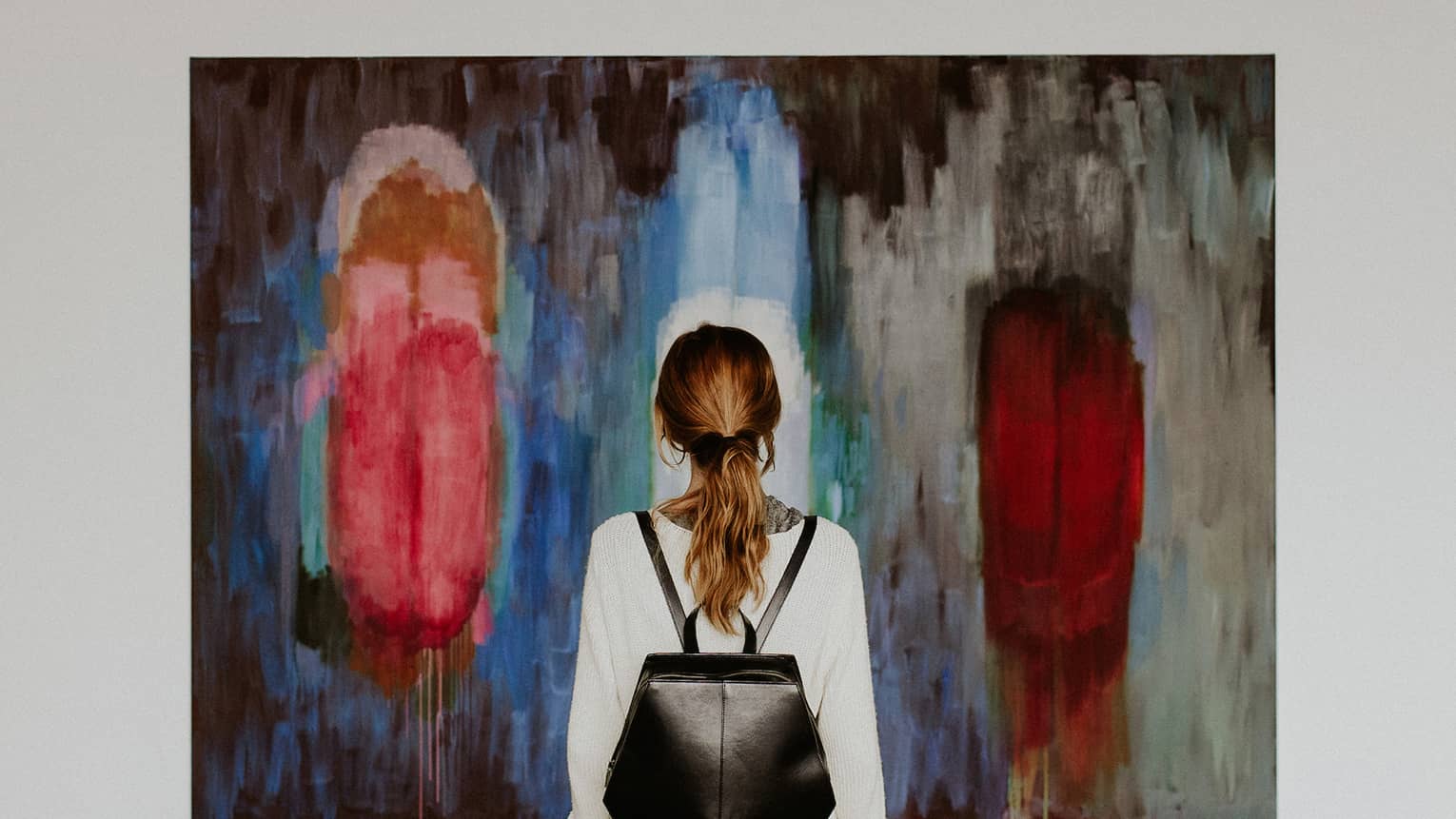 A person with a backpack standing in front of an abstract painting in an art gallery.