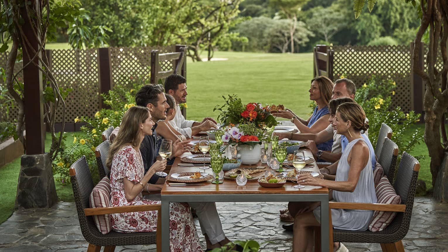 Eight people sit around a long dining table set up outside beneath a canopy of greenery and string lights