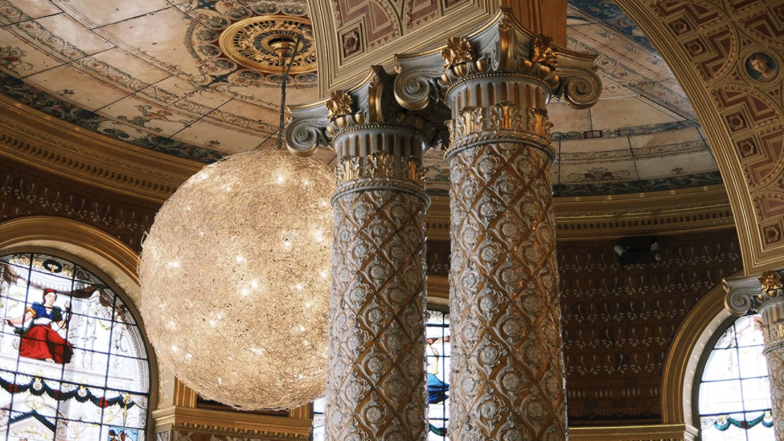 An interior space with a high arches and columns with decorative inlay and carving. Stained-glass windows, decorative ceiling and walls, and chandelier lighting are in the background.