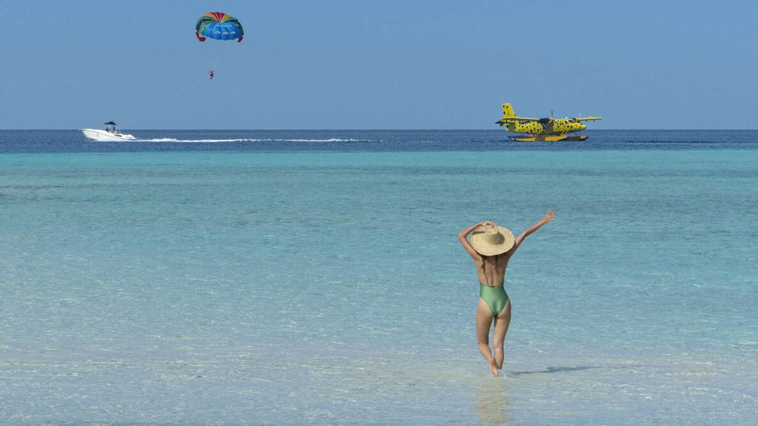 A woman walking on the beach watches a seaplane and a parasailor in the distance. 