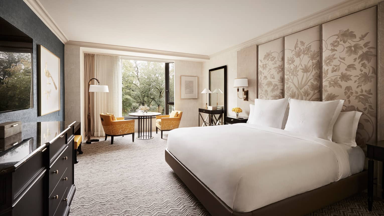 Deluxe Garden-View Room white bed with tall fabric floral headboard, black dresser with TV, yellow velvet chairs by window