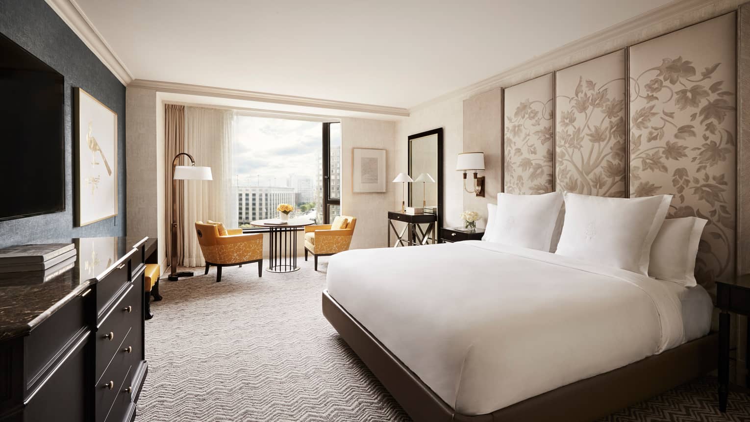 Deluxe City-View Room white bed with tall floral fabric headboard, black dresser, yellow velvet dining chairs by window