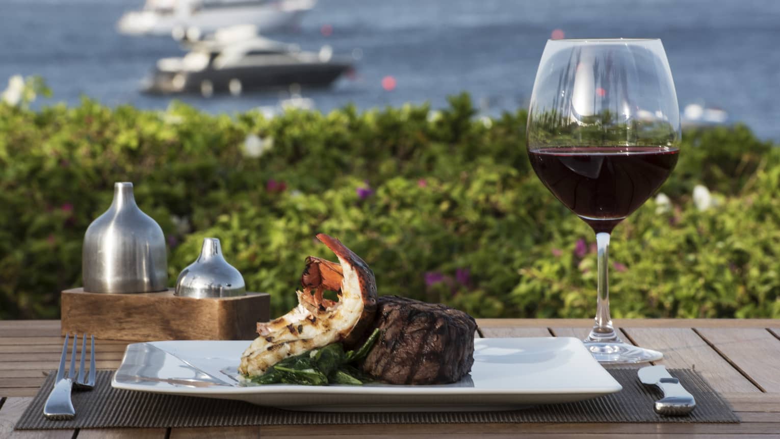 Plate with grilled lobster tail, steak and red wine on patio table overlooking sea, boats